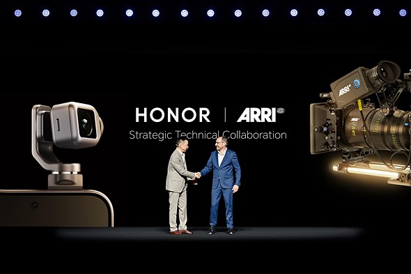two men in suits stand shaking hands on a stage in front of a screen with a smartphone and large cinema camera