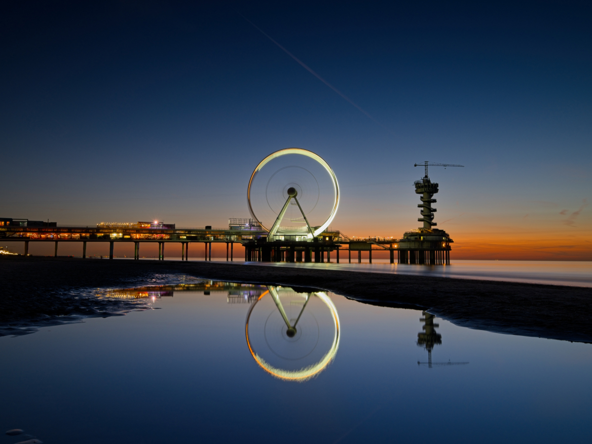Creative urban photography of a Ferris wheel at dusk, perfectly reflected in still water with a glowing circular symmetry against a deep blue sky.