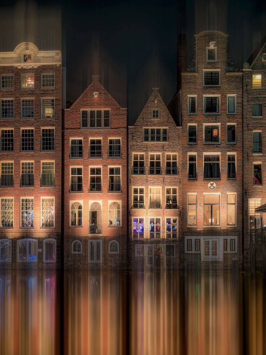 Urban reflection photography of historic canal houses at night, with warm glowing windows mirrored in the water and stretched into a soft vertical reflection effect.