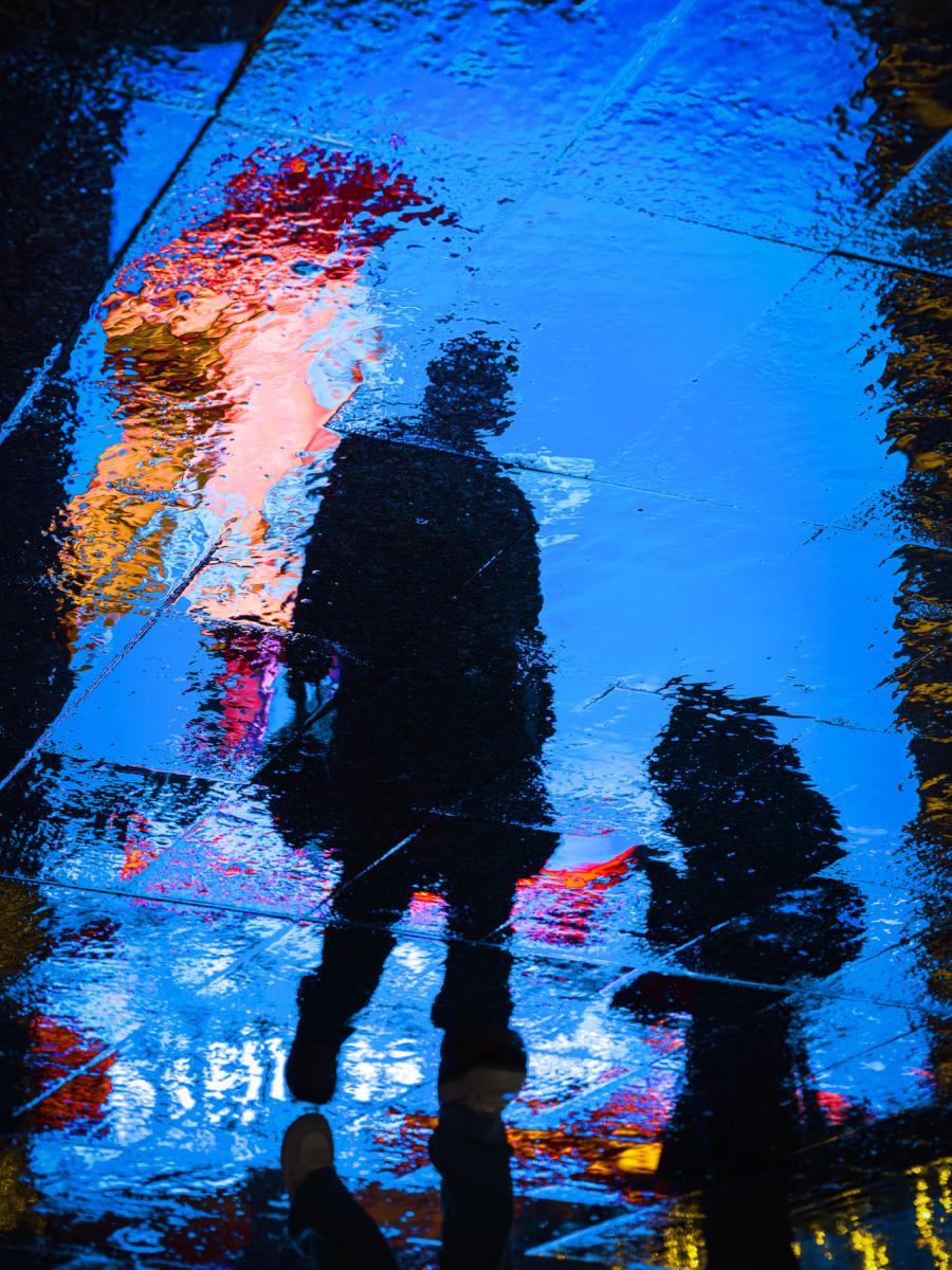 Creative urban photography showing a person and dog reflected in a rain-covered street, with vivid blue tones and abstract light reflections creating a surreal city scene.