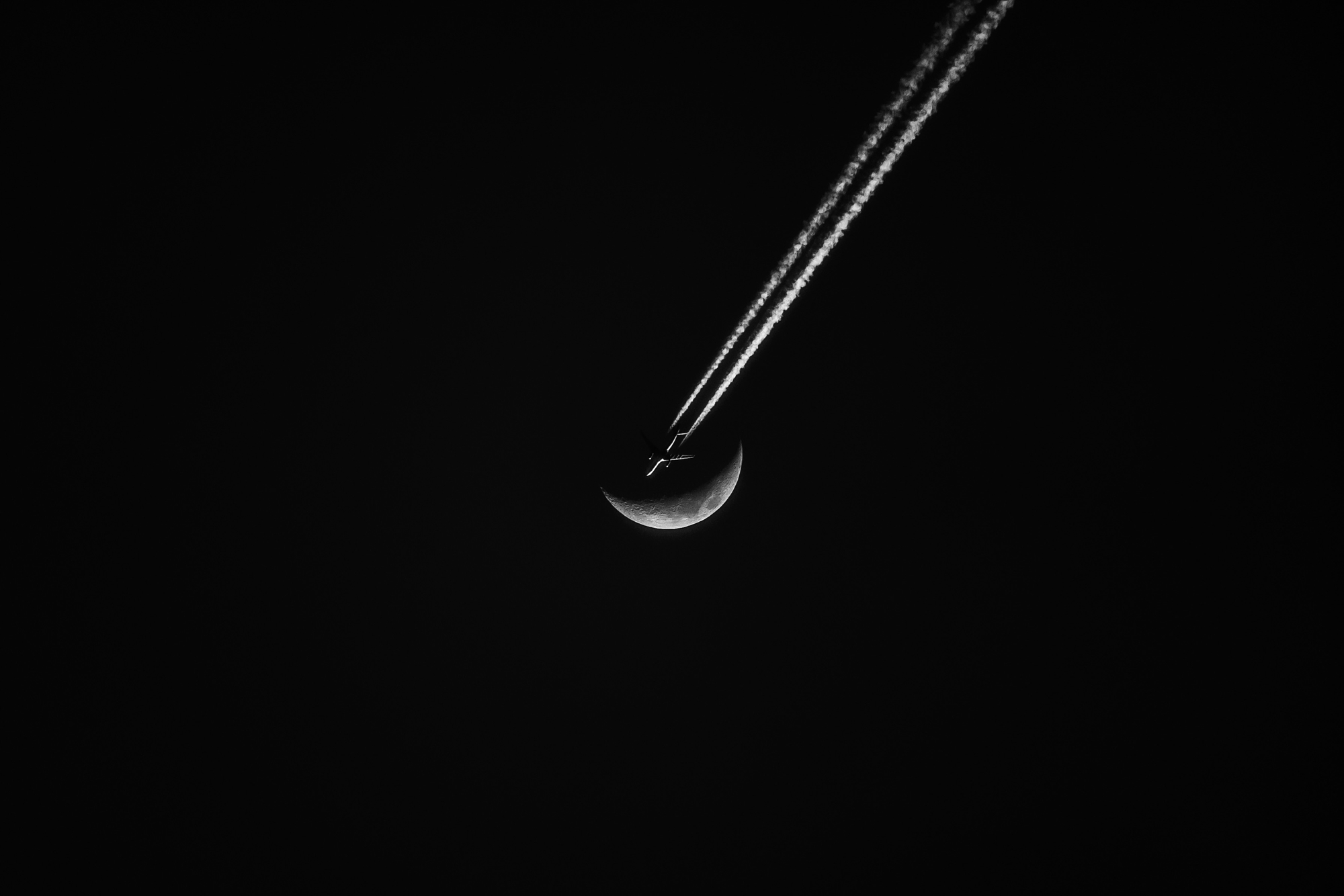 David Baca black and white photograph of an airplane crossing a crescent moon with visible contrails