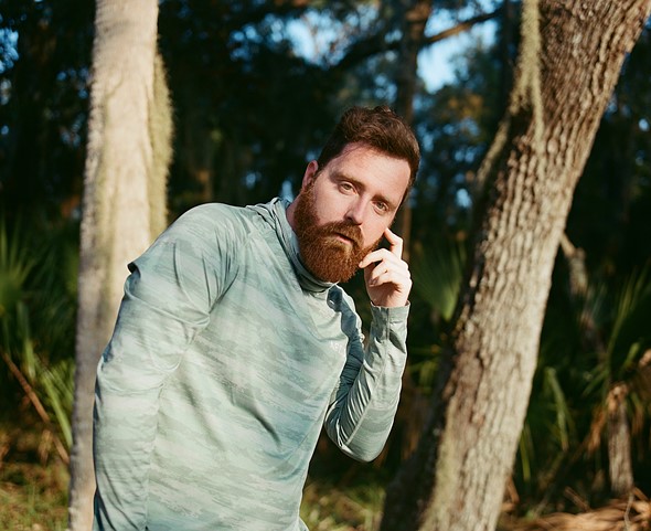 A bearded man in a sage green athletic pullover leans forward among trees  resting his fingers against his cheek and looking directly at the camera
