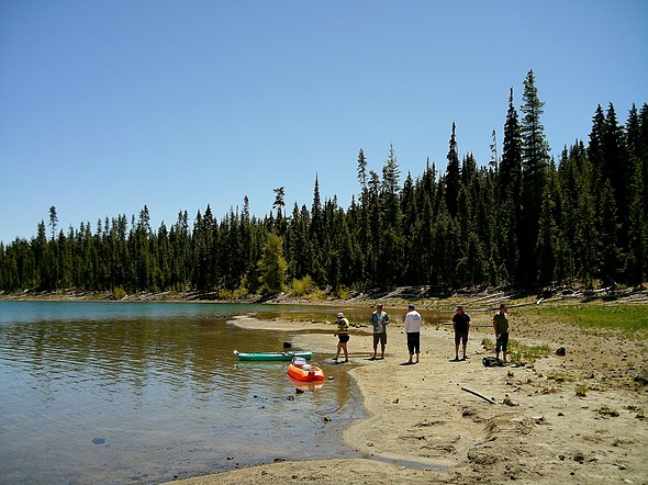 Four people on a sandy lake shore with a green and an orange kayak in shallow water