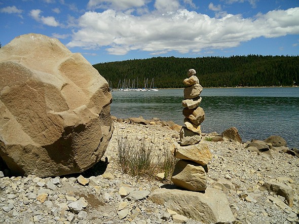 Stack of balanced rocks on a rocky lakeshore near a large boulder  with calm water  forested hills  and sailboats in the background