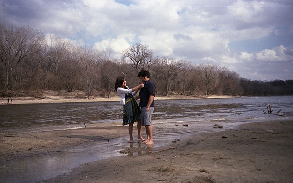 a couple stands barefoot on the sandy banks of a river with bare trees behind them