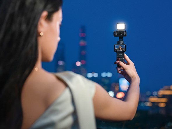 a woman holds a small camera gimbal with light at arms reach with a city nightscape behind