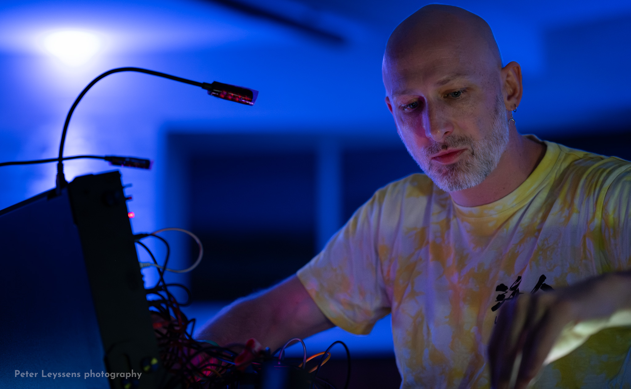 Individual in a yellow shirt adjusts electronic equipment with visible wires and a small microphone, under blue and purple lighting; “Peter Leyssens photography” text appears in the corner.