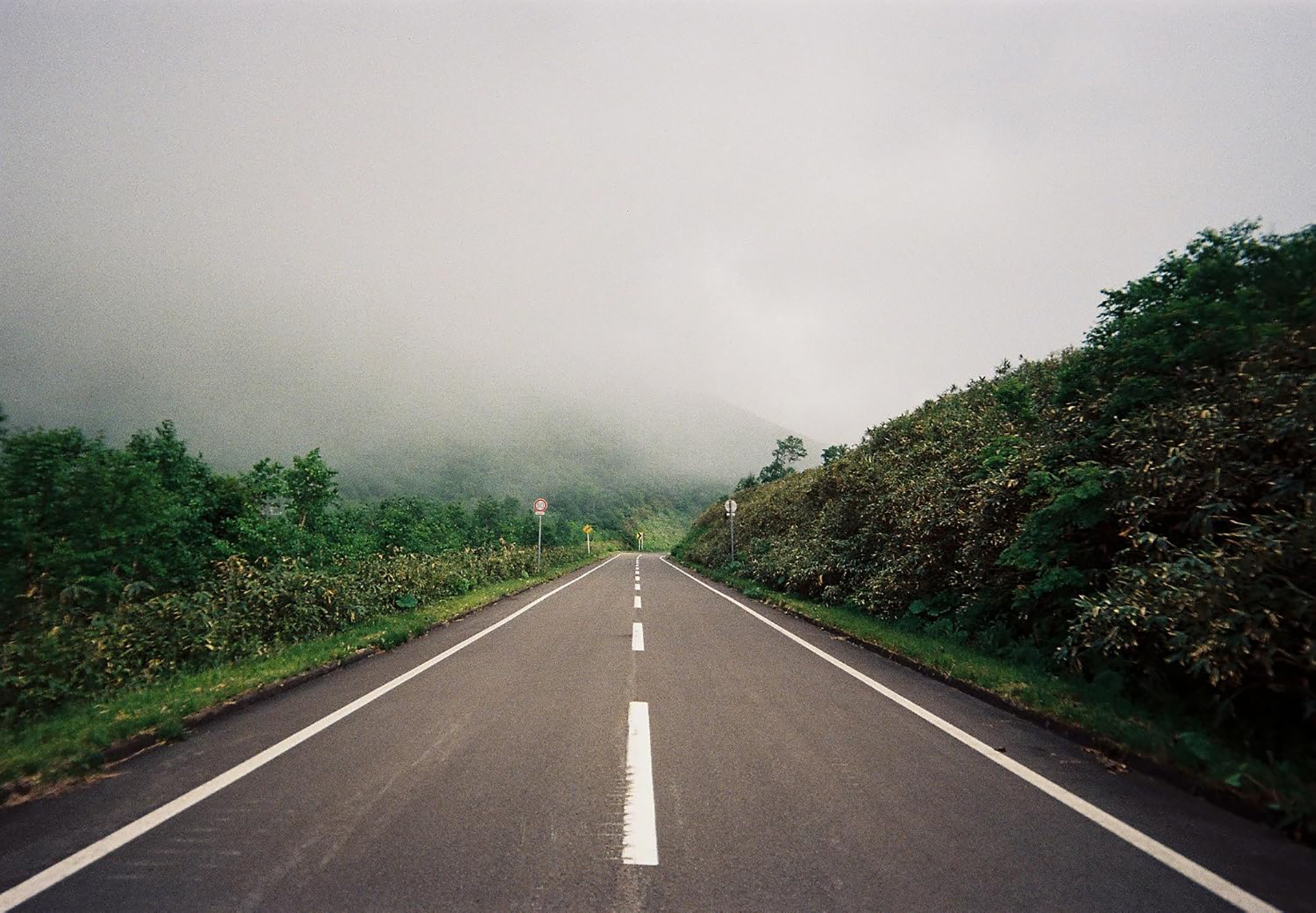 An empty road extends into the distance, bordered by green bushes and trees, with misty hills visible under a cloudy sky. Scene photographed using a KODAK EC35 reusable 35mm film camera.