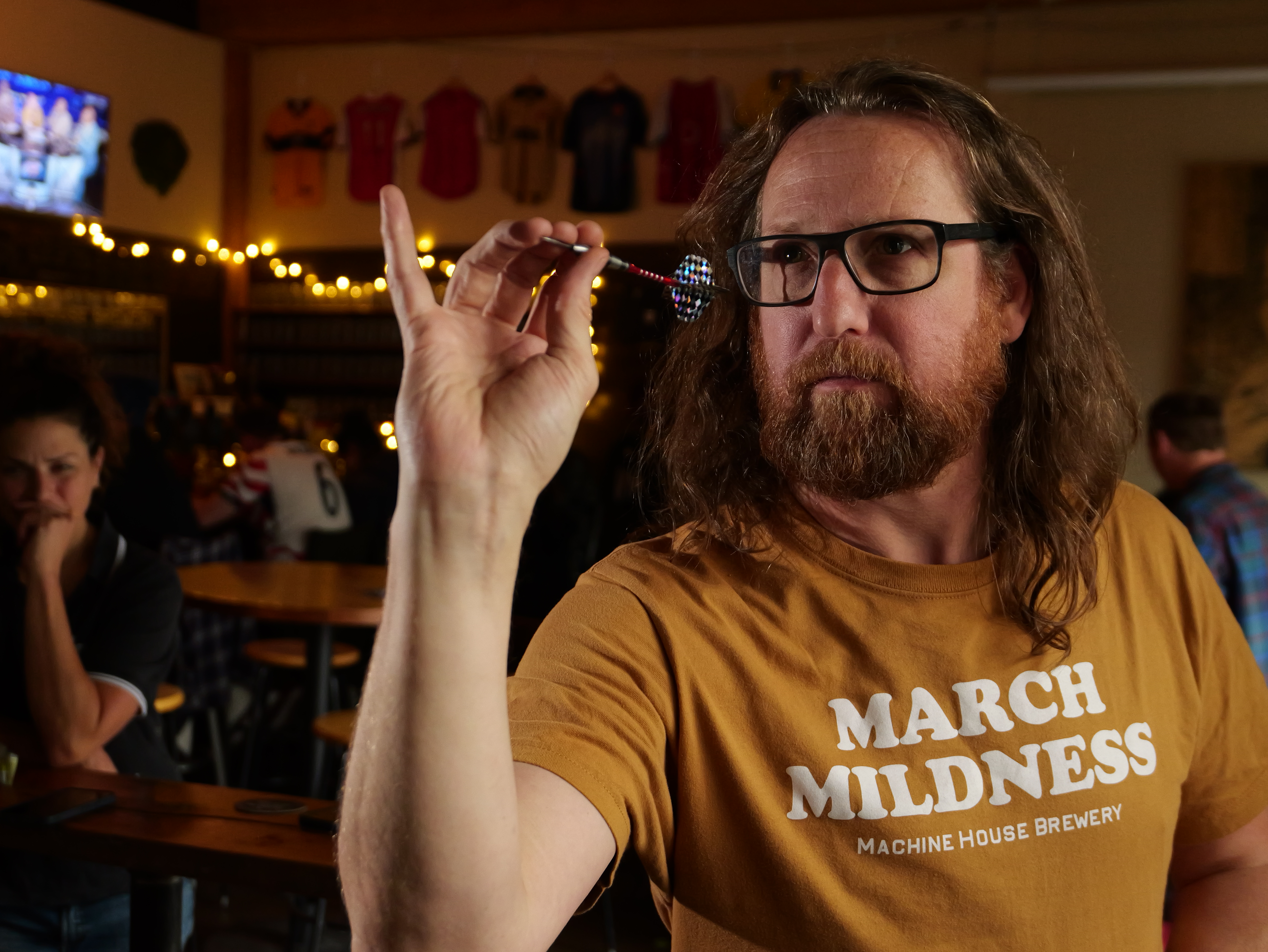 A man with flowing hair and a beard holds a dart and looks intently off to the side of the image