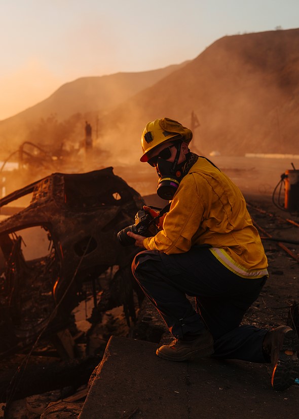 a person wearing a respirator helmet and nomex hacjet kneels down holding a camera amidst charred vehicles and smoke