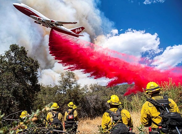 firefighters stand watching as a plane drops red fire retardant over a forest with smoke behind it