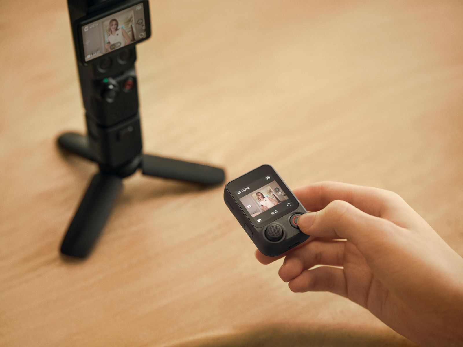 A photo of the DJI Osmo Pocket 4P on a table, with a hand in front of it holding the FrameTap remote, a small module with a screen and controls.