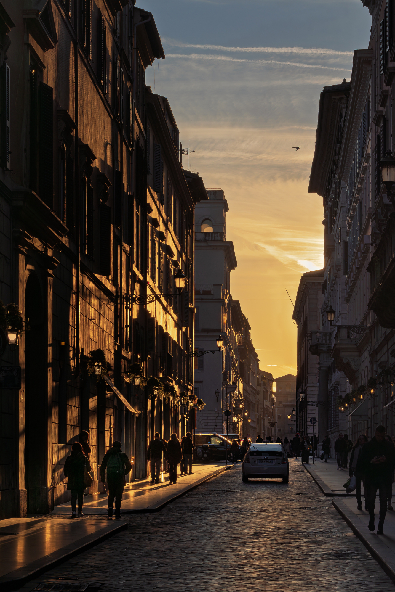 Pedestrians walking down a narrow European cobblestone street illuminated by the warm, directional glow of golden hour photography.