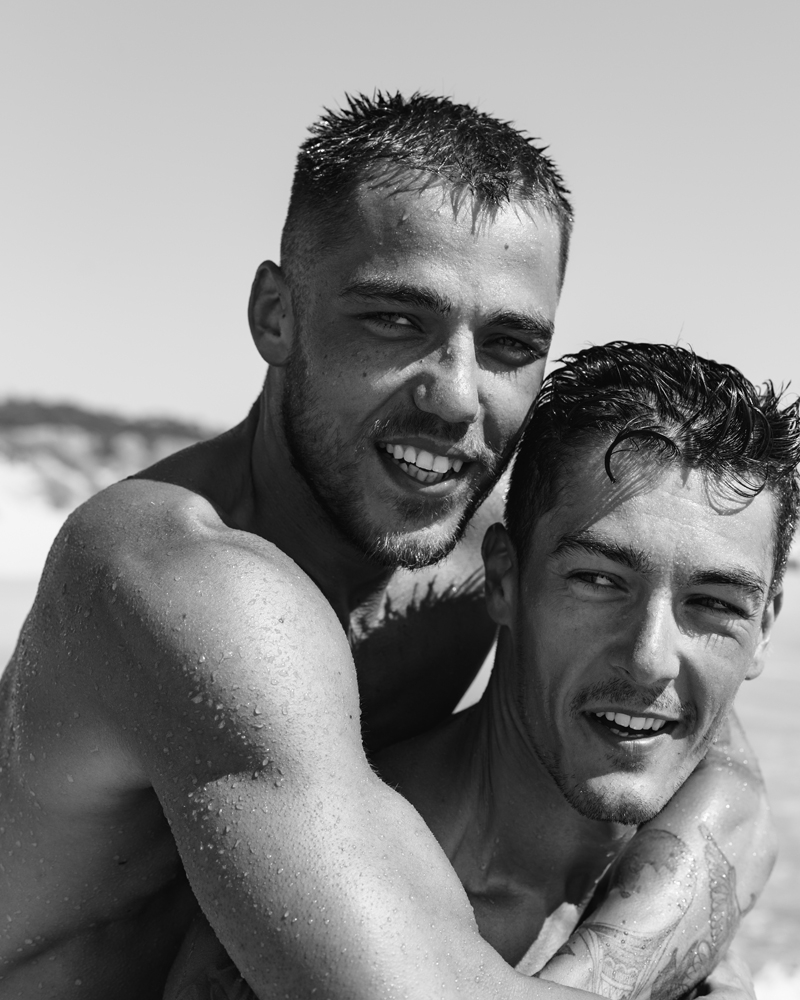 Black and white portrait of two smiling men embracing on a beach, highlighting raw emotion and male beauty photography by David Velez.