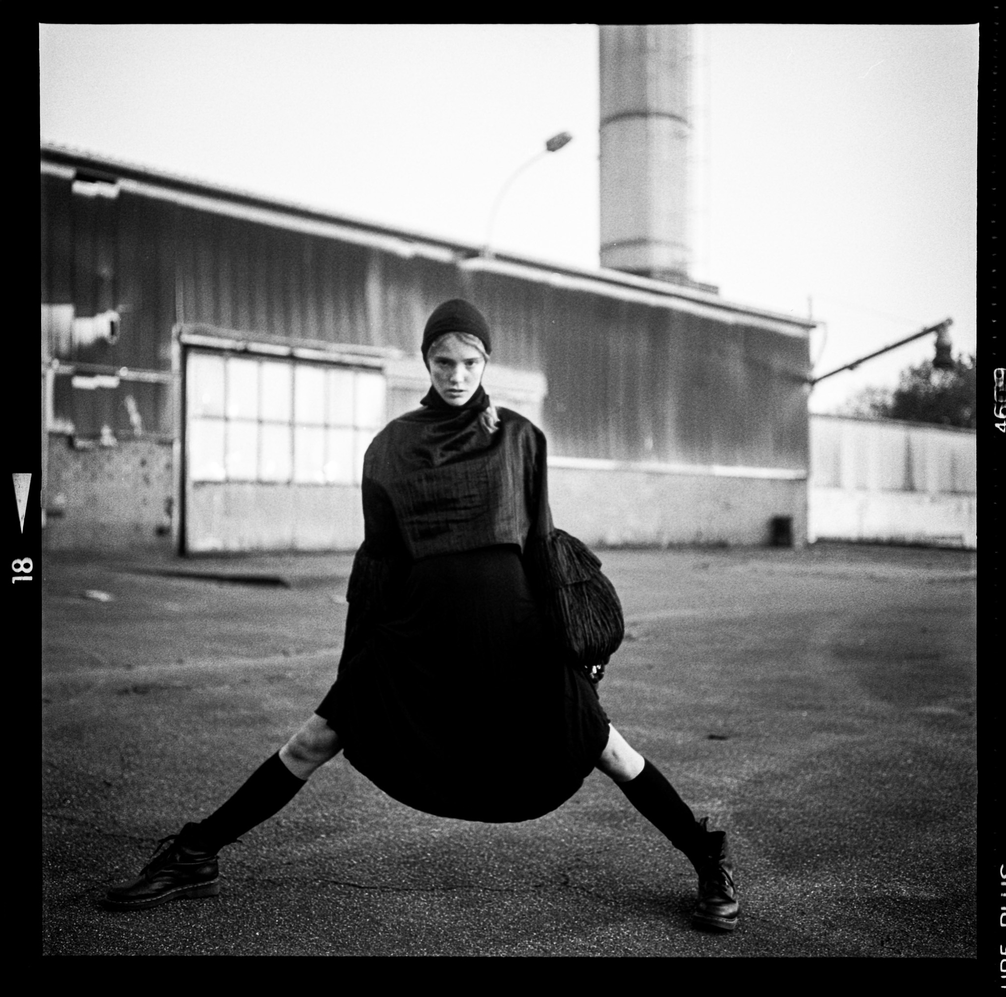 Medium format black and white fashion photograph by Holger Nitschke of a model posing in dark attire outside an industrial building.