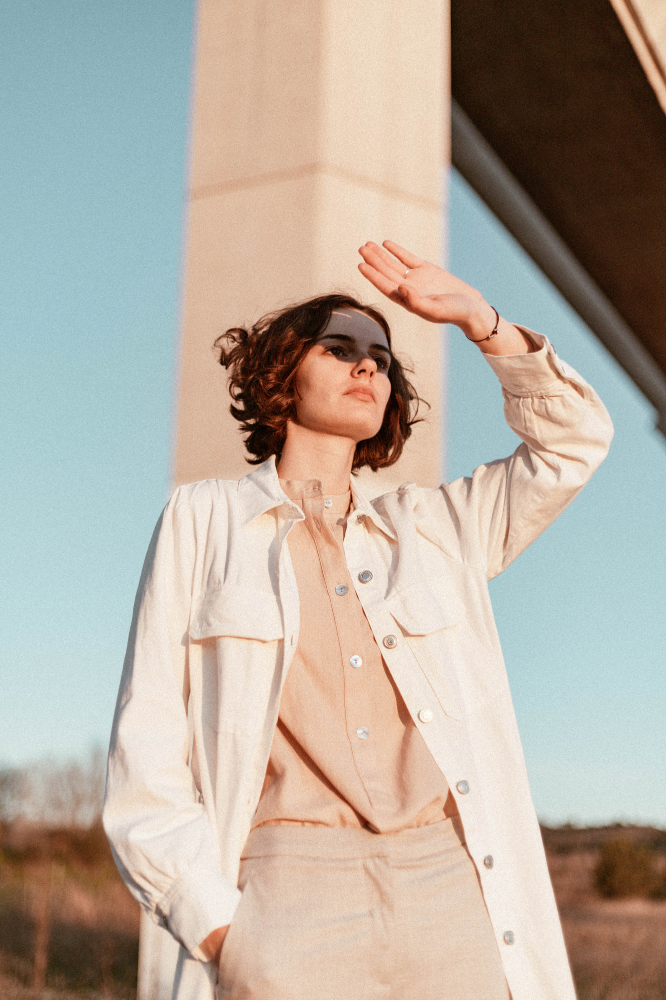 Portrait of a woman standing near a bridge shielding her eyes from bright midday light, showcasing harsh light photography techniques and open shade setups.