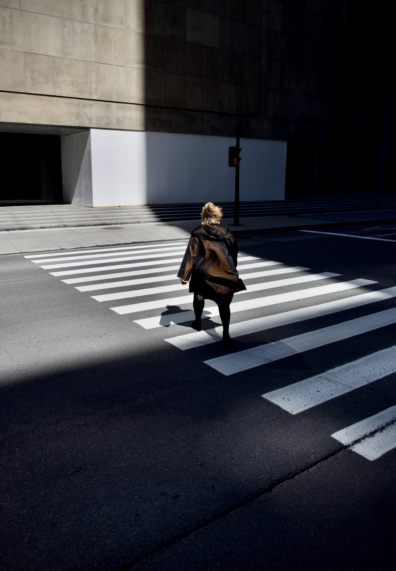 Street photography lighting demonstration of a pedestrian walking across a crosswalk with dramatic high contrast light and deep geometric shadows on urban pavement.