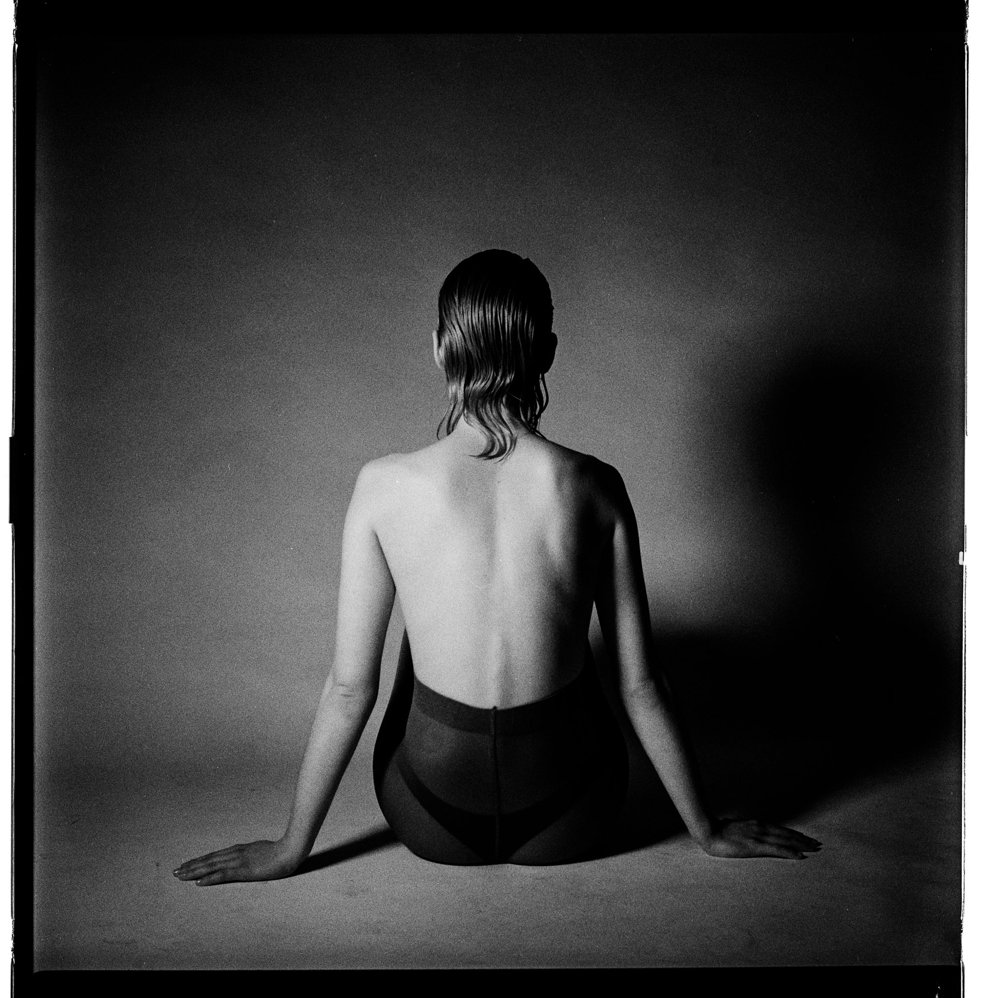 Minimalist studio black and white photography by Holger Nitschke showing the rear view of a seated woman in light and shadow.