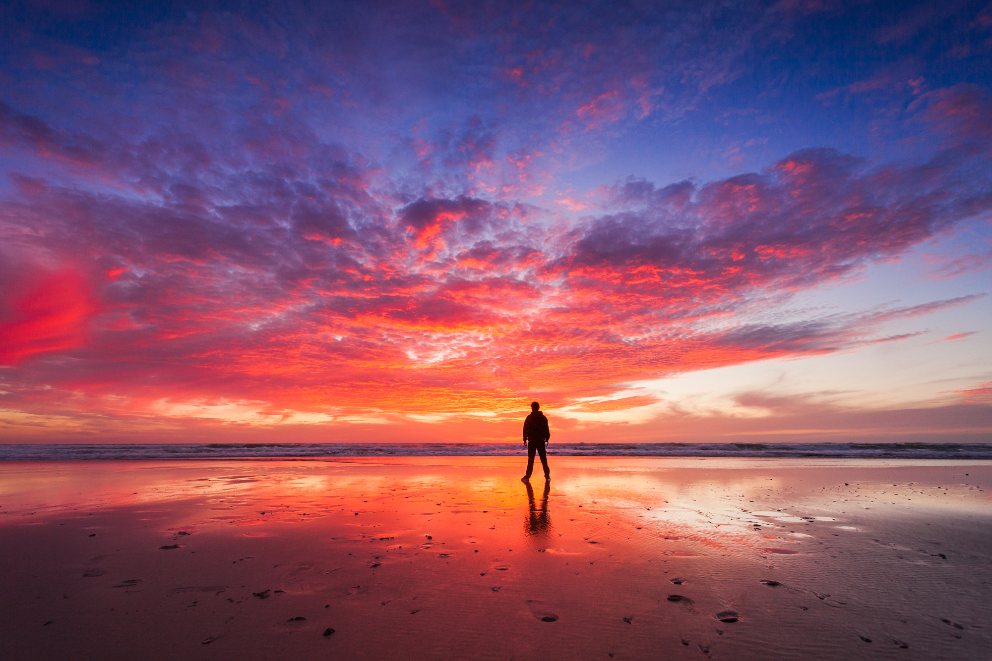 Silhouette of a person standing on a reflecting wet beach beneath a vibrant pink and orange sunset sky, a powerful example of Younes Ezzentri landscape photography.