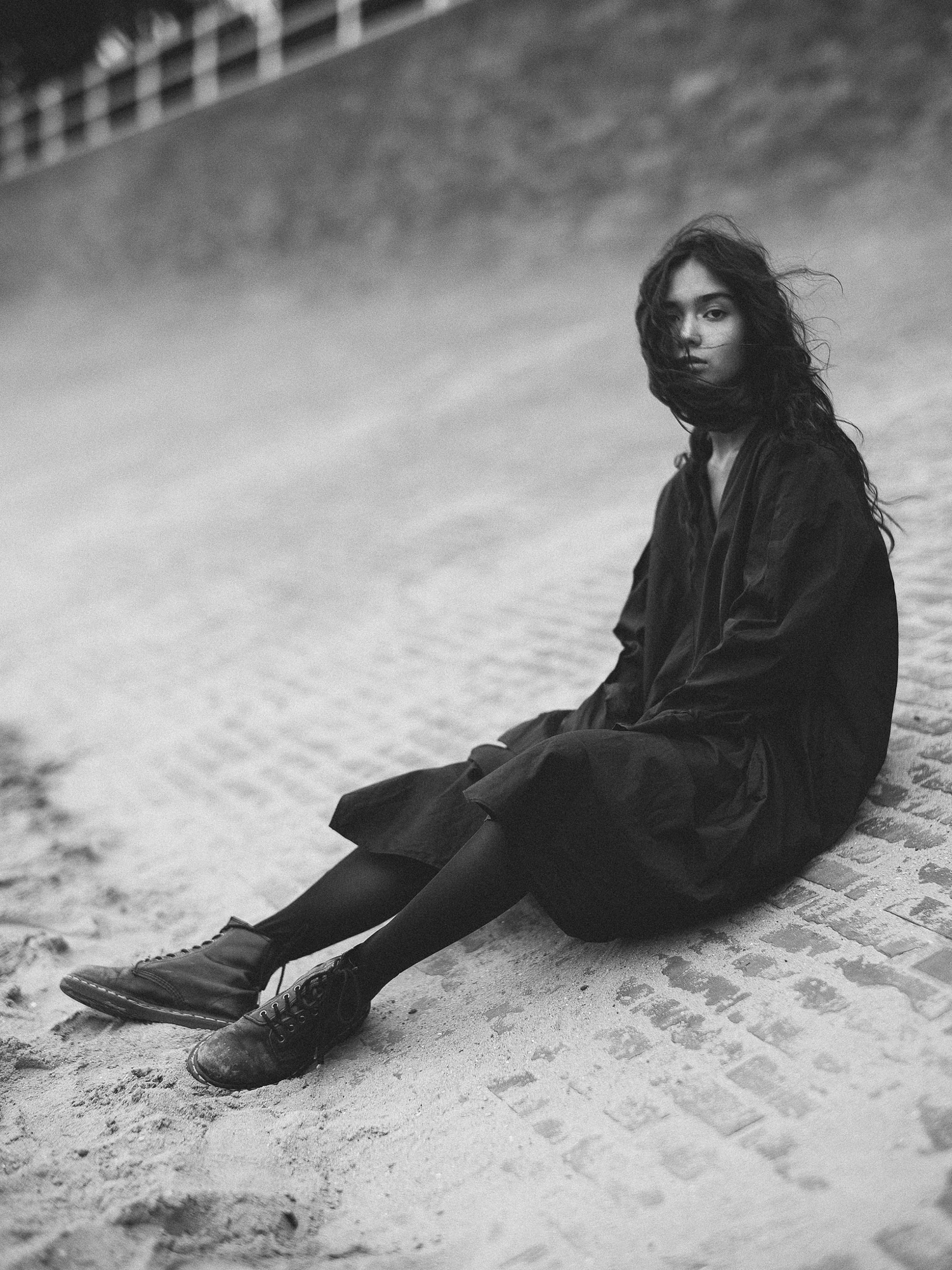 Mood-driven outdoor black and white portrait by Holger Nitschke of a woman in a dark dress sitting on a sandy slope.