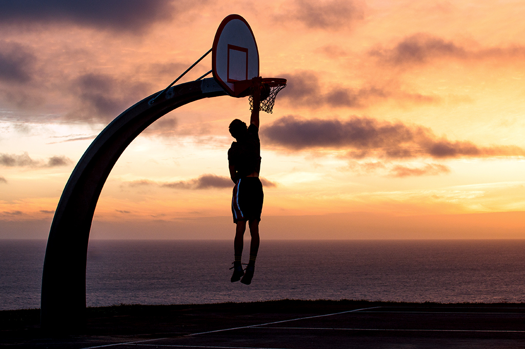 Sunset silhouette photography of a basketball player hanging from a hoop against a golden sky over the ocean.