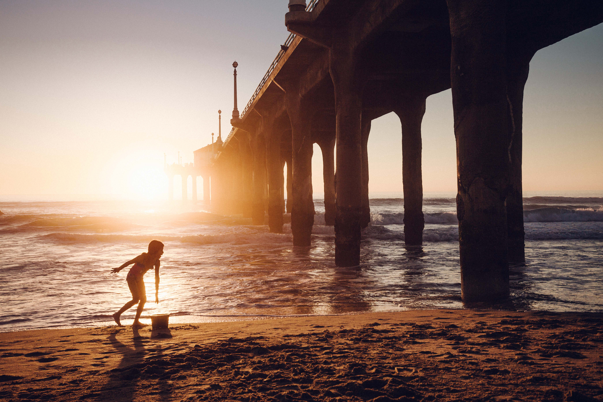 Sunset silhouette photography of a child playing on the beach beneath a large ocean pier.