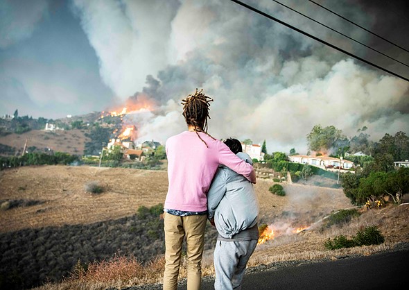 two people stand in an embrace looking out at a large fire engulfing a hillside with houses
