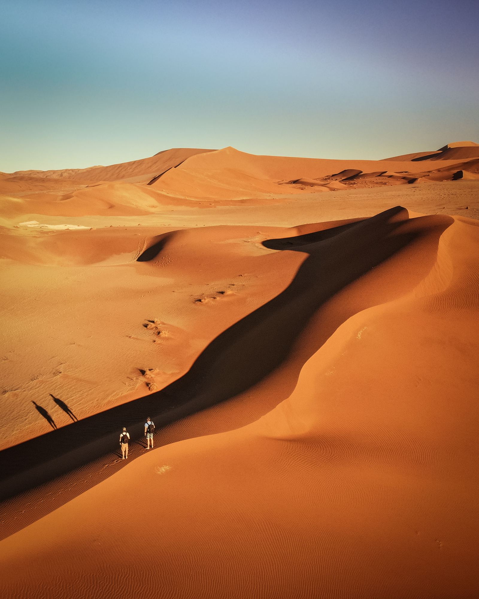 High-angle view of two hikers walking through orange desert sand dunes under a soft blue sky, demonstrating teal and orange photography contrast.