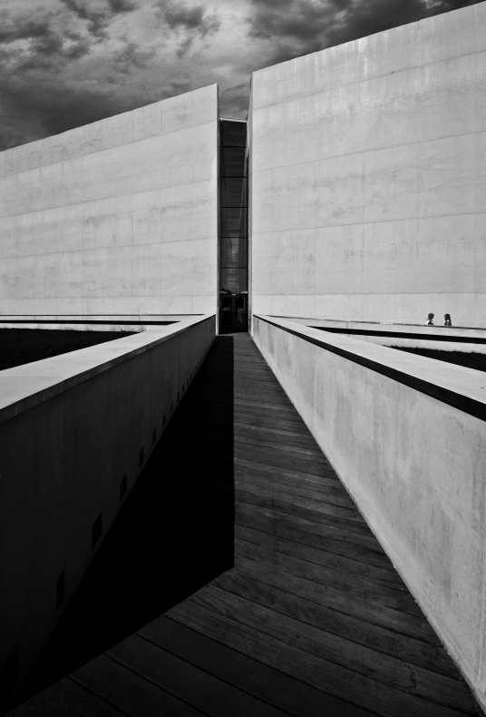 High-contrast black and white view of a wooden walkway between two tall concrete walls. Two small figures stand at the far end under a stormy sky.
