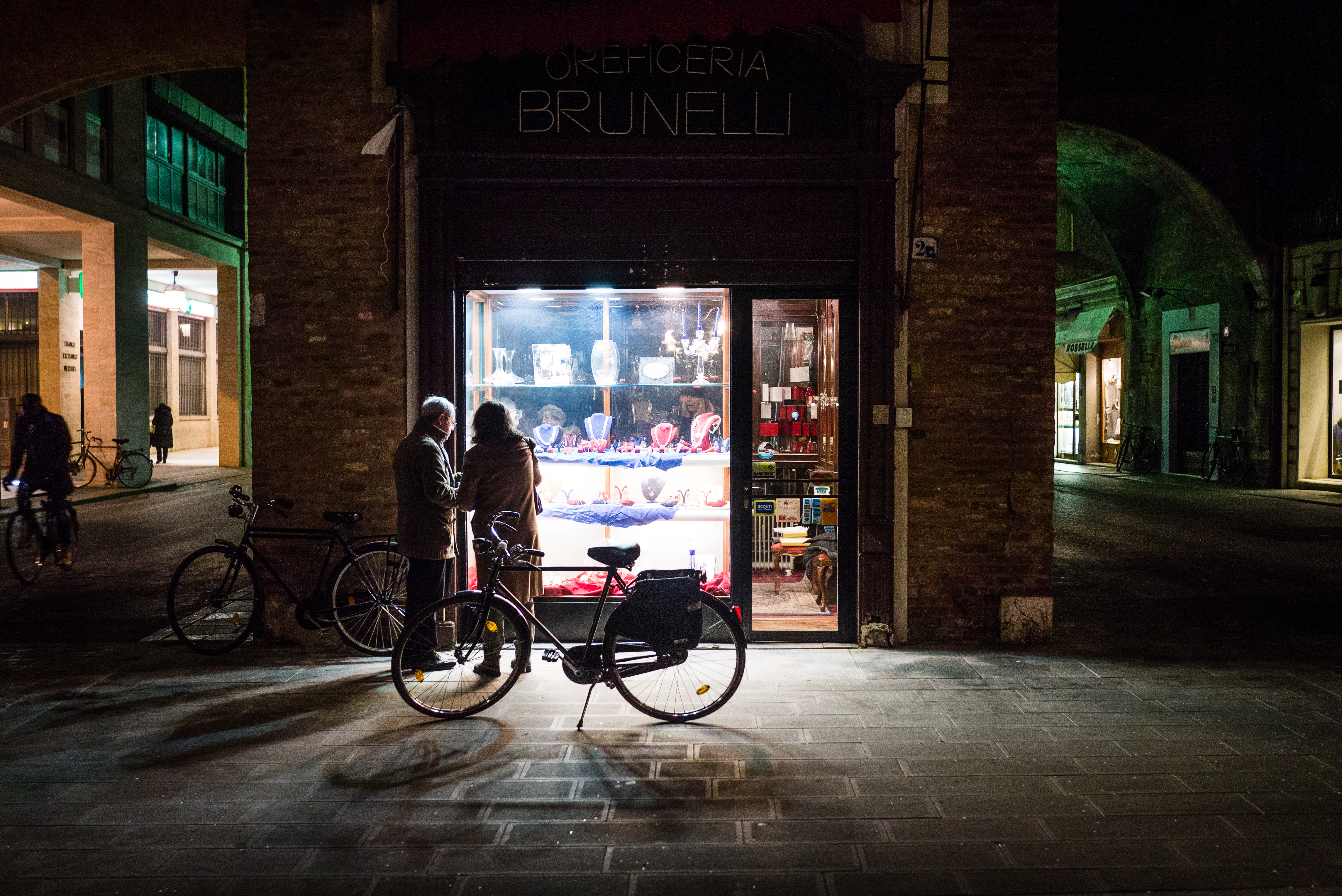 Everyday storytelling photography: an older couple looks into a lit jewelry shop window at night, a bicycle parked in front.