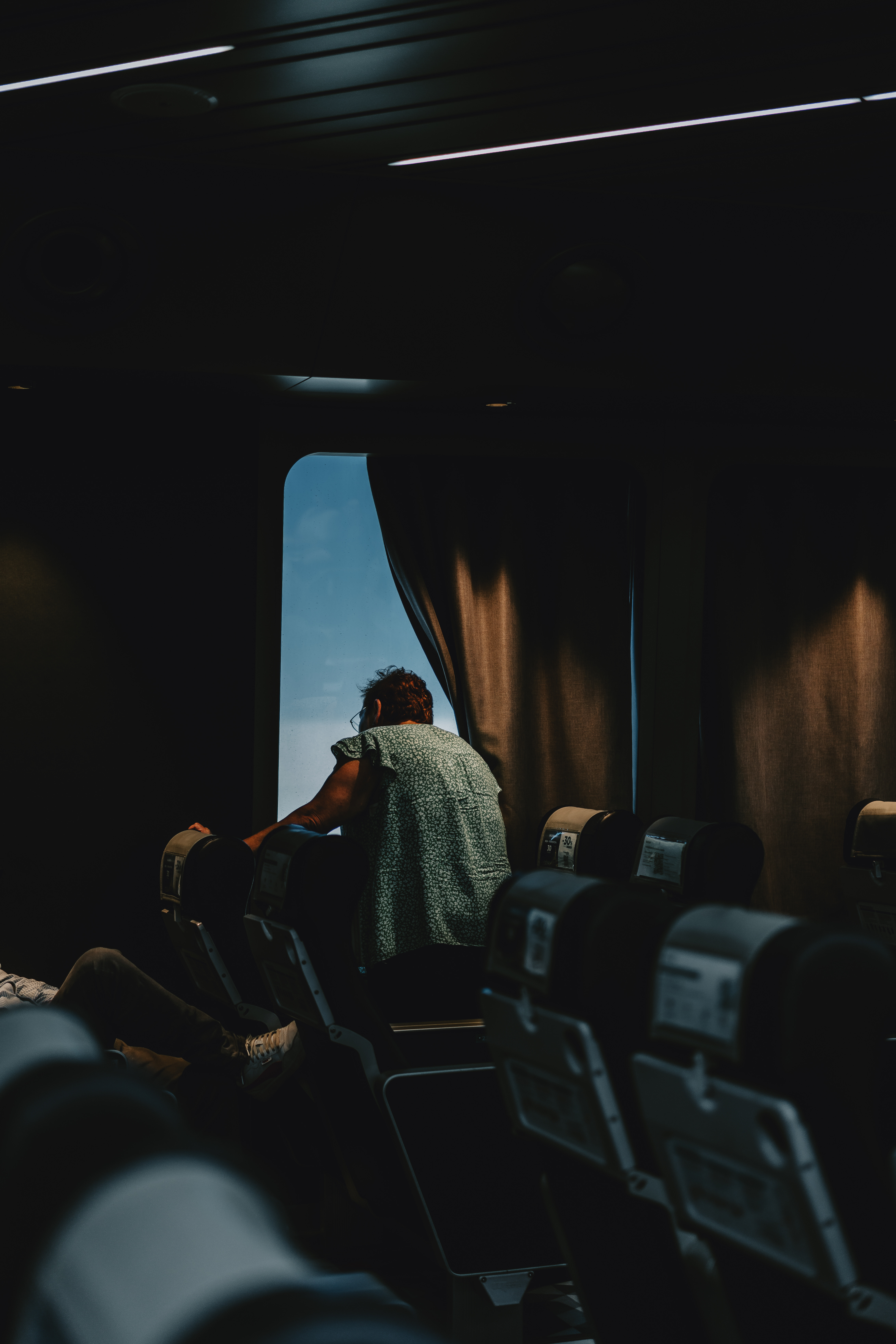 Passenger leaning past a curtain to look out a bright train window in a darkened carriage, from Eline Deckers's street photography.