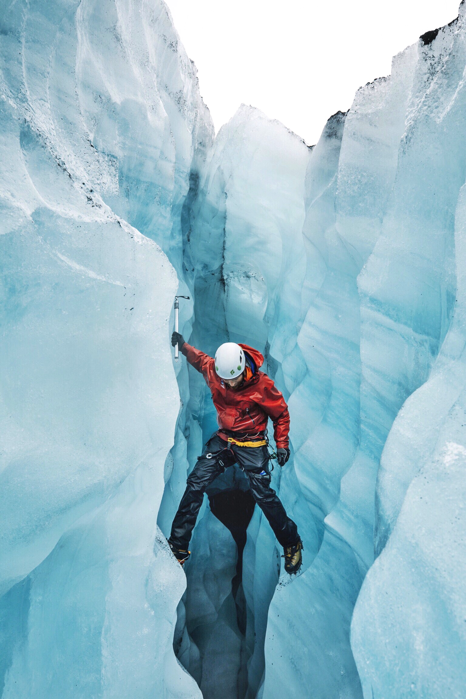 Ice climber in a red jacket ascending a pale blue glacier crevasse, a warm and cool tones photography contrast that heightens tension.