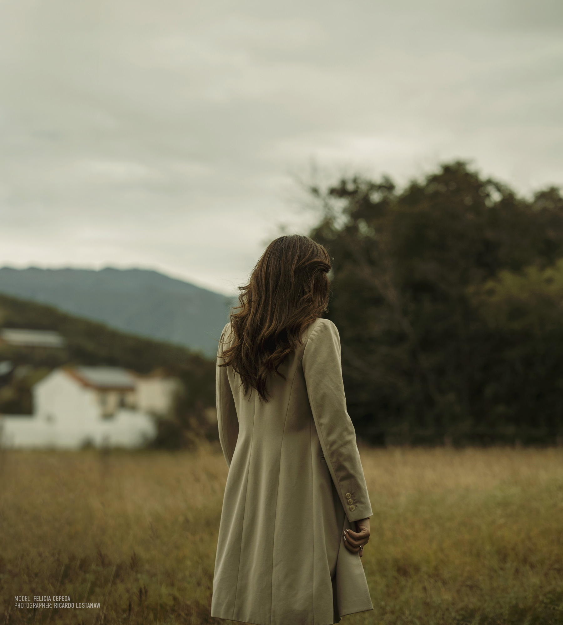 Woman in a pale coat photographed from behind in a field below the mountains