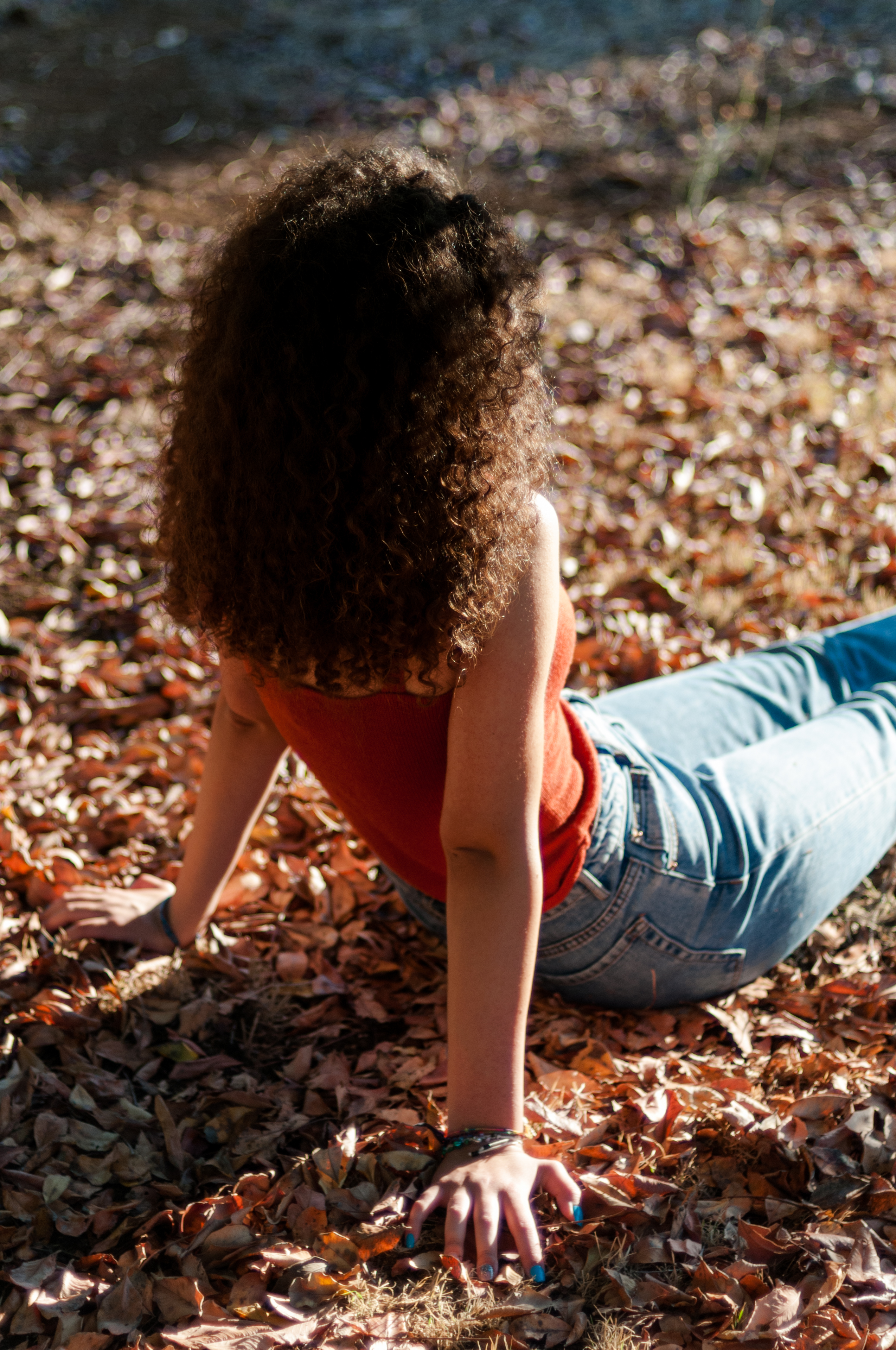 Backlit woman with curly hair, a red top and jeans sitting in fallen autumn leaves, an example of people in fall foliage photography.