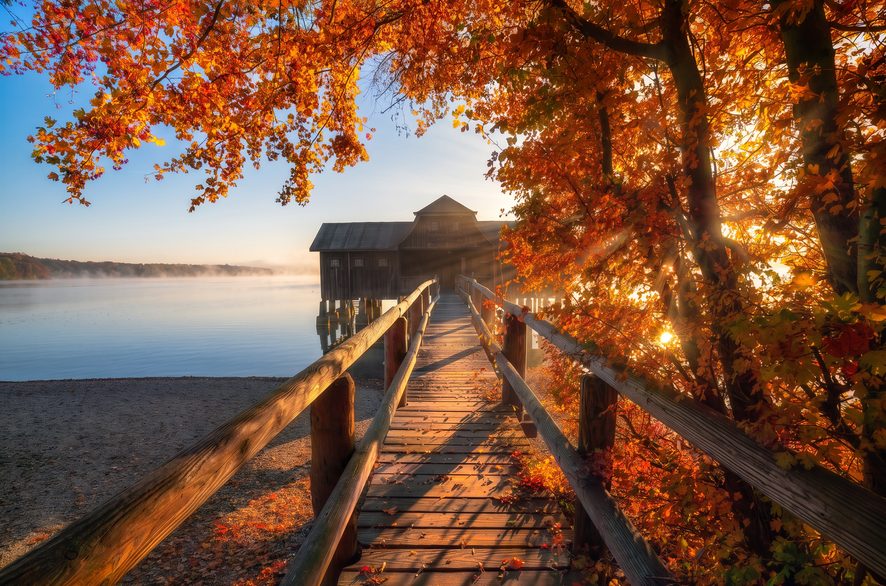 Wooden boardwalk leading to a boathouse on a misty lake, framed by backlit orange leaves with the sun bursting through the branches.